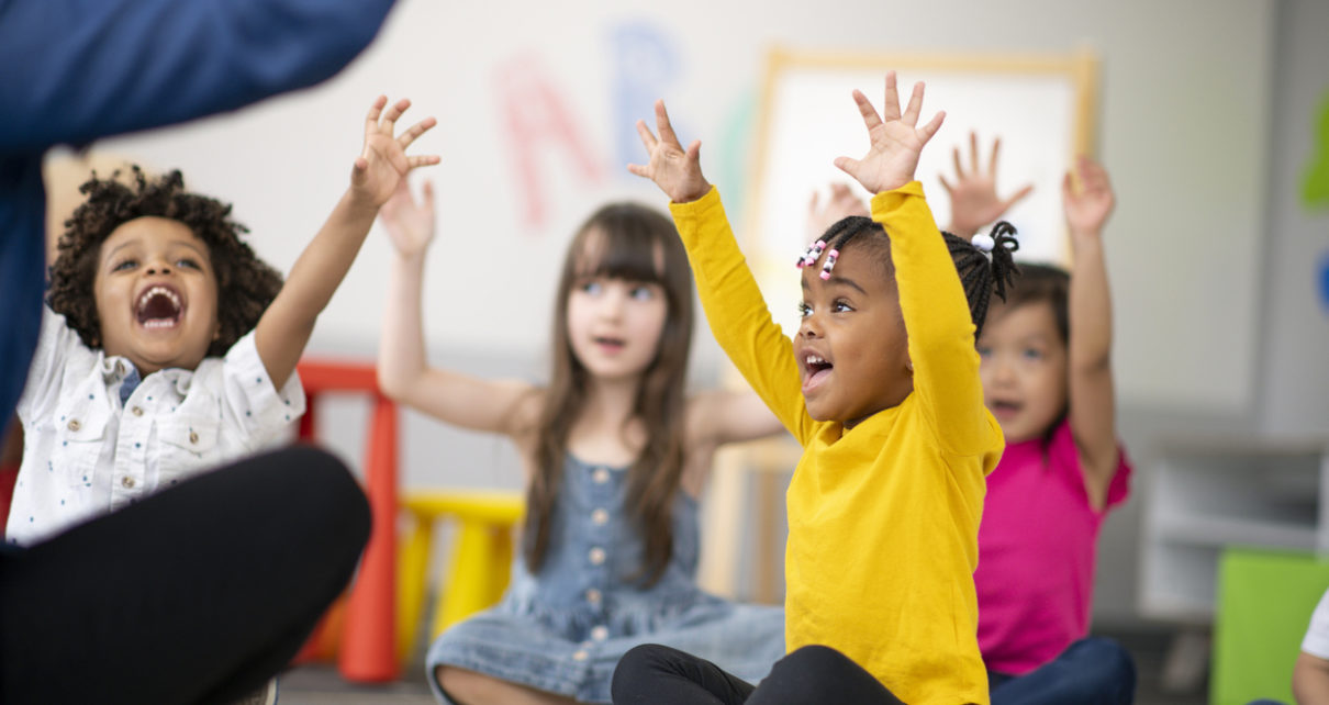 A multi-ethnic group of preschool students is sitting with their legs crossed on the floor in their classroom. The mixed-race female teacher is sitting on the floor facing the children. The happy kids are smiling and following the teacher's instructions. They have their arms raised in the air.